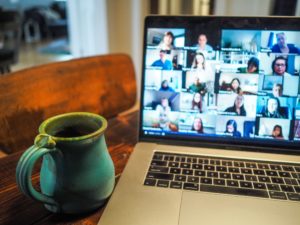 Image of a laptop screen with a video call taking place, displaying several people in small boxes on the screen. A coffee cup is to the left of the laptop.